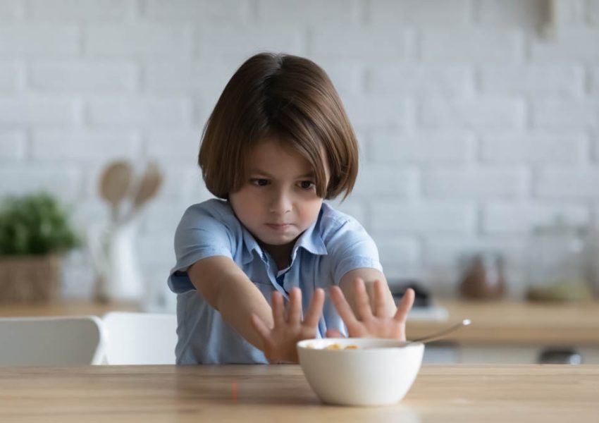 Stubborn little kid boy rejecting eating dry breakfast corn flakes with milk, sitting in kitchen without appetite. Upset small male child does not want eating tasteless food, loss of appetite.